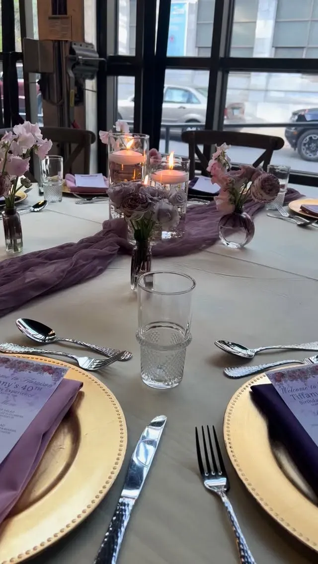 Candlelit event table with delicate pink and mauve floral arrangements in bud vases, gold chargers, and soft draped linen.